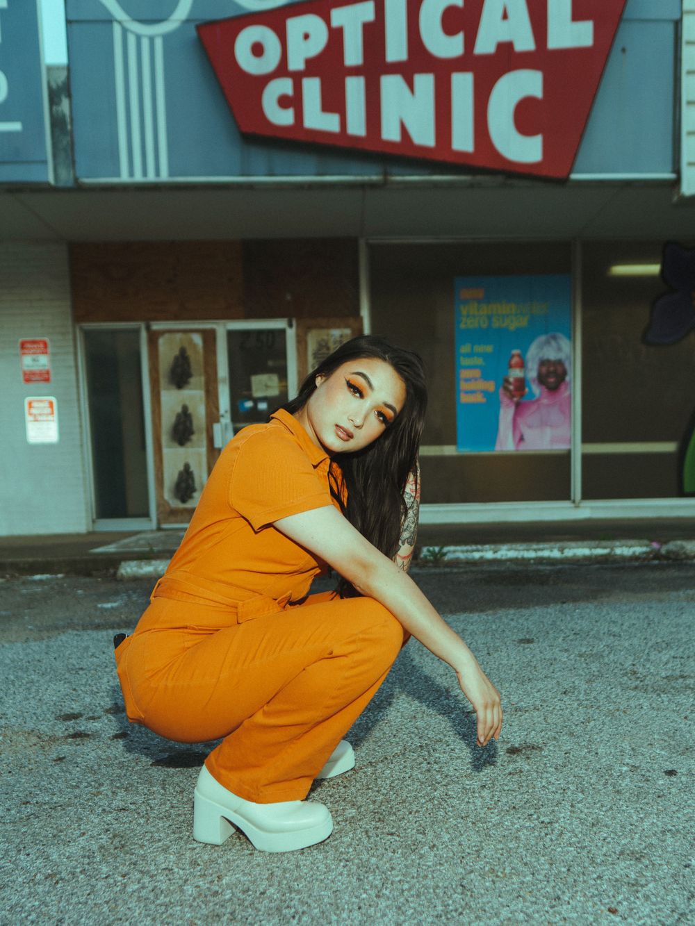 Fashion model crouches in orange jumpsuit and white platforms outside vintage optical store.