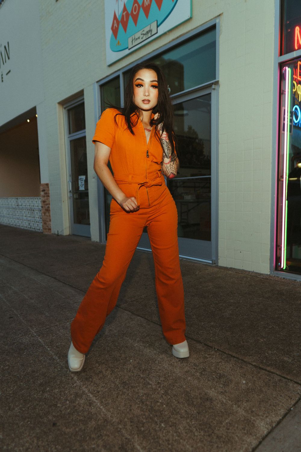 Stylish portrait of model in vibrant orange jumpsuit against urban storefront backdrop.