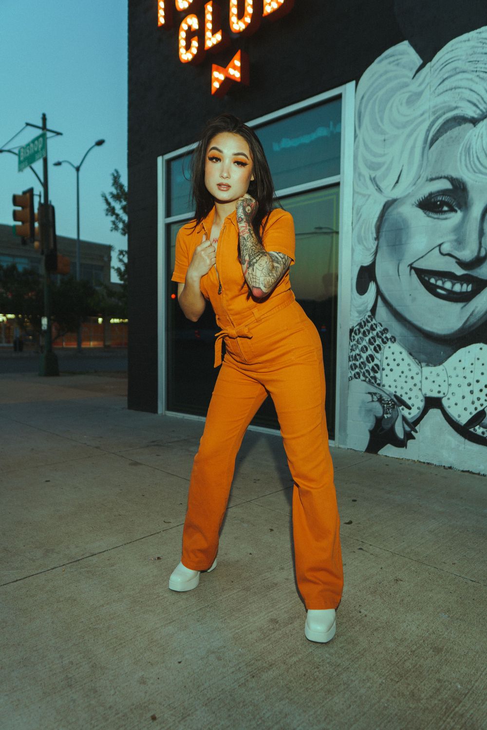Orange jumpsuit fashion shoot against urban wall with Dolly Parton mural visible in background.