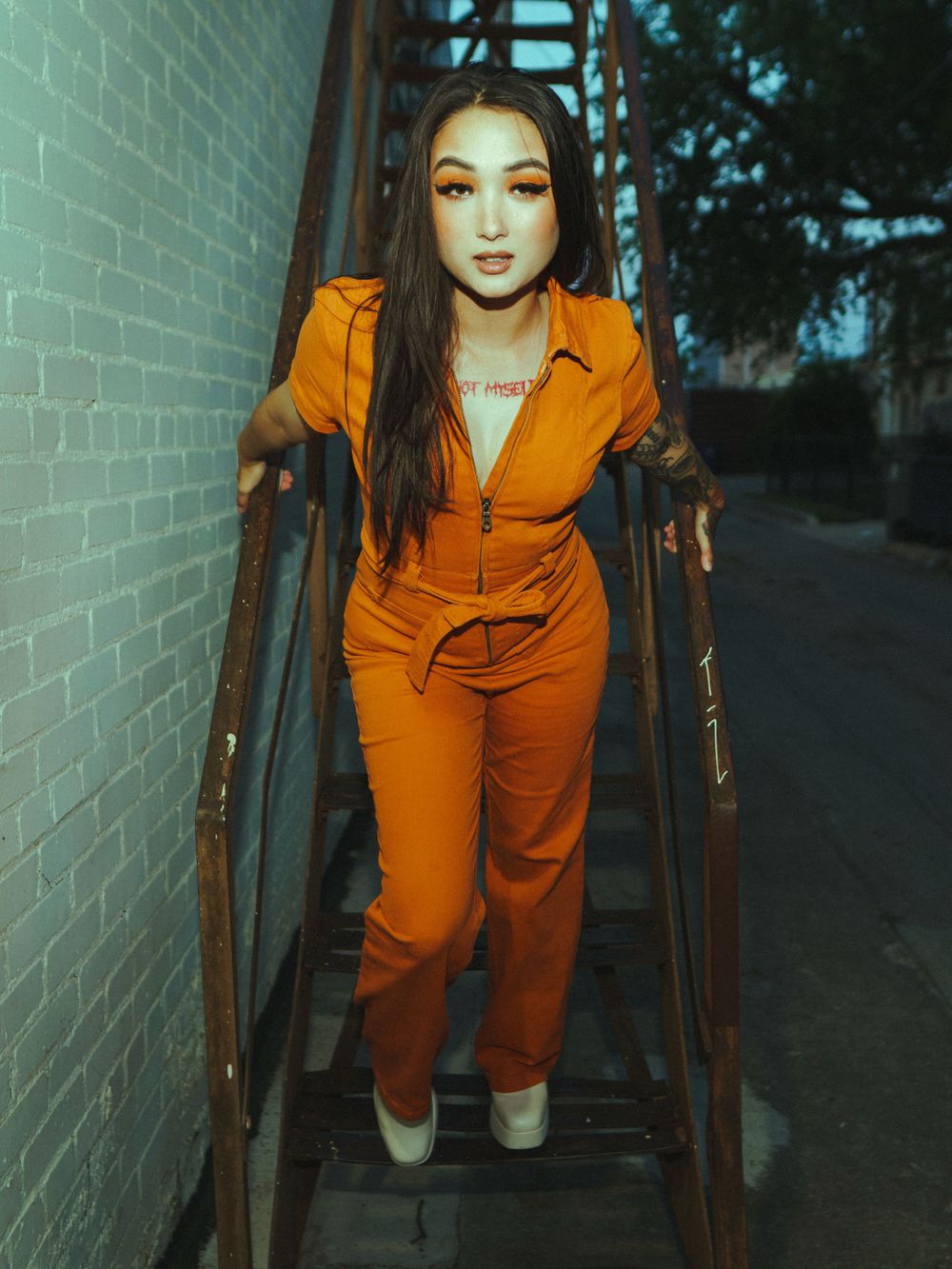 Model descends metal stairs in fitted orange jumpsuit against moody urban backdrop.