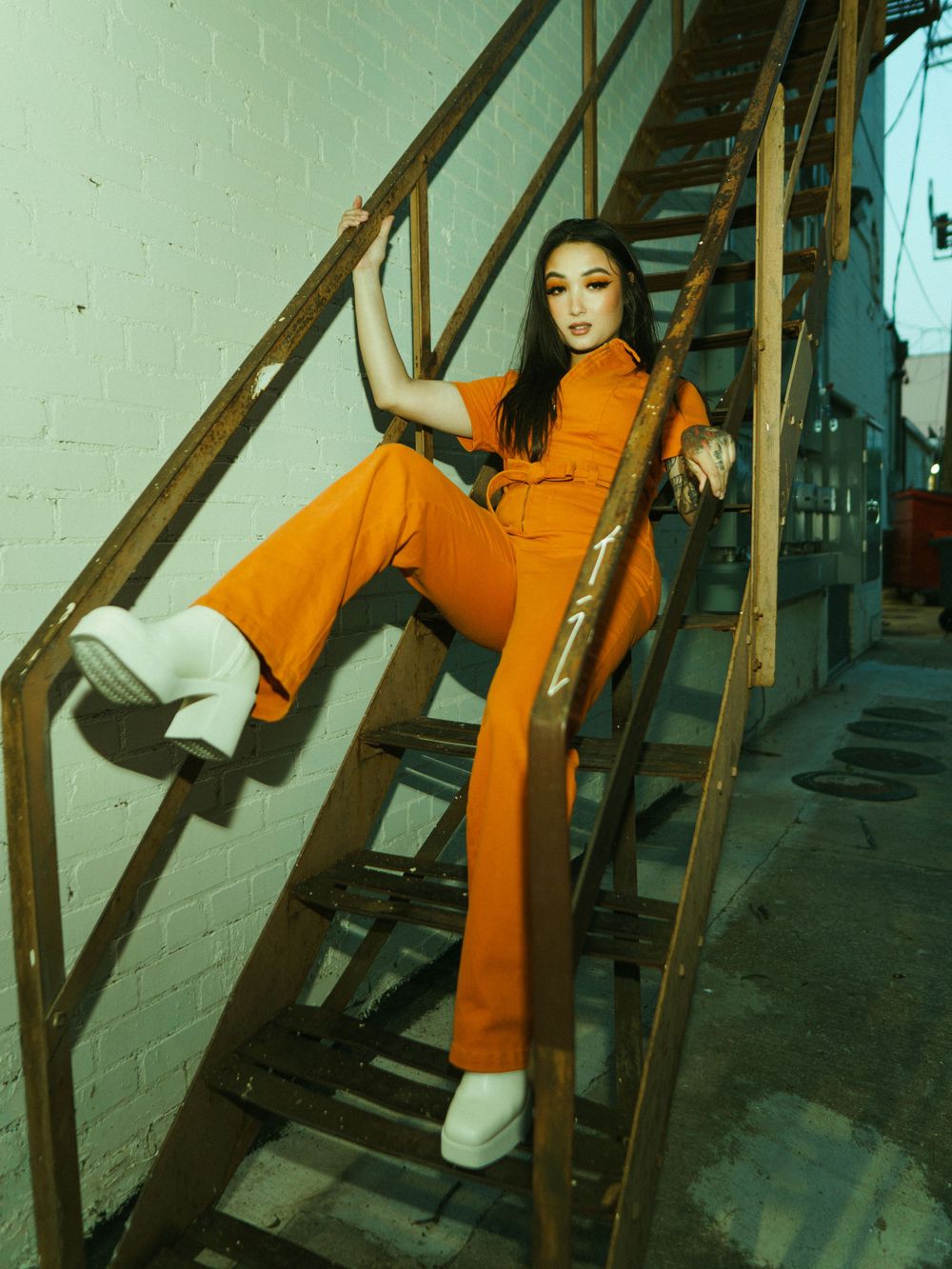 Model poses dramatically on industrial metal staircase in orange utility jumpsuit.