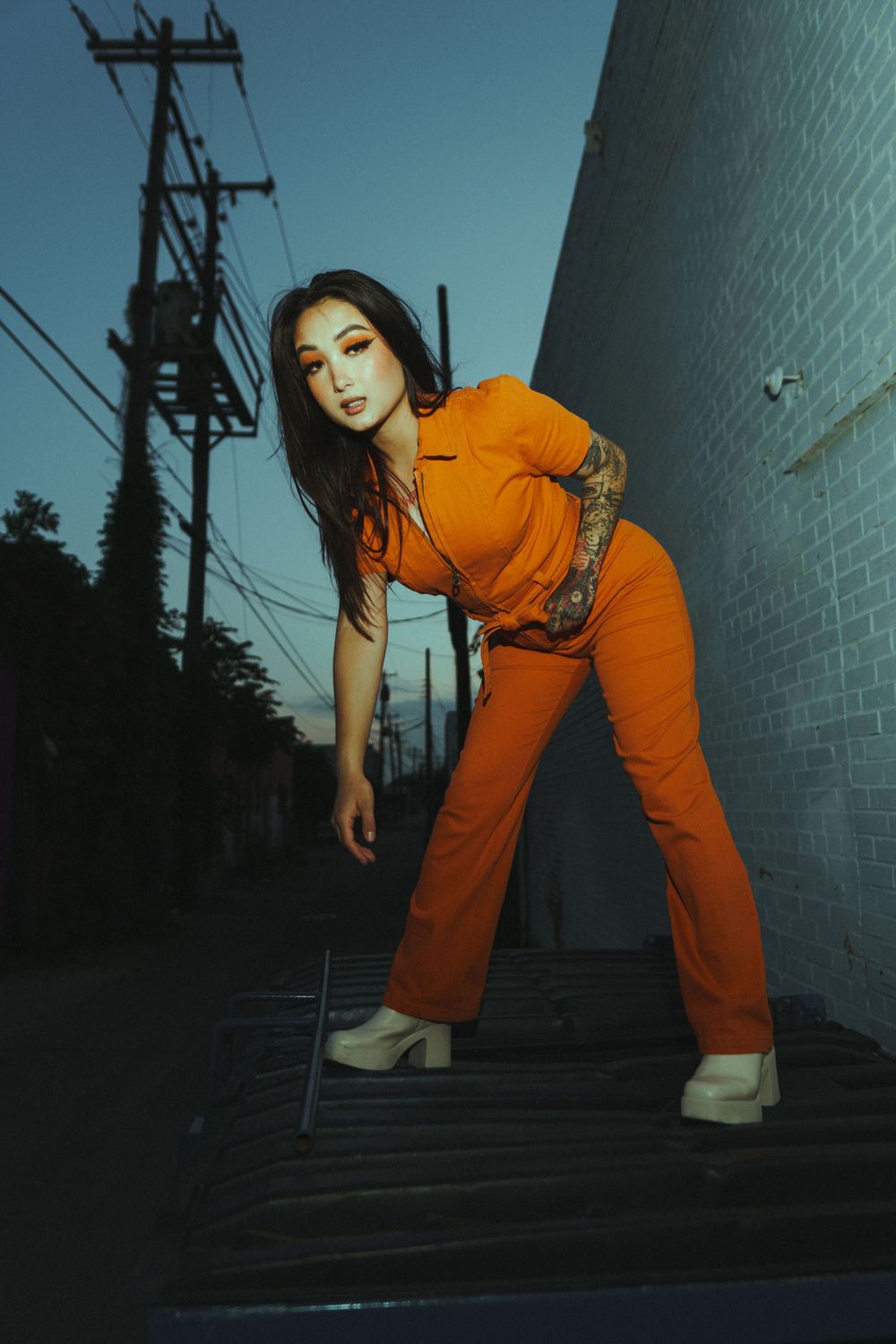 Fashion model poses on metal stairs at twilight in vibrant orange utility jumpsuit.