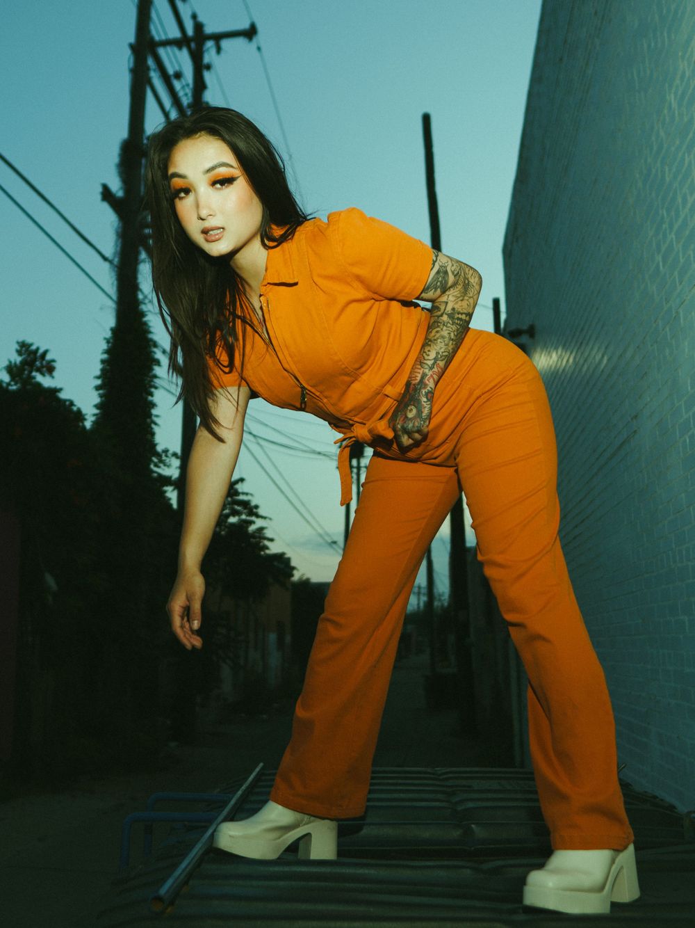 Dramatic dusk photoshoot featuring orange jumpsuit against industrial urban backdrop.