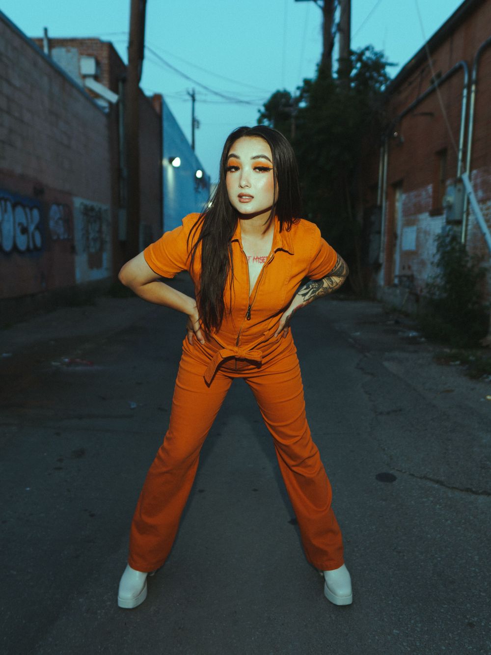 Fashion photo in an urban alley featuring a bright orange jumpsuit against a moody blue evening backdrop.