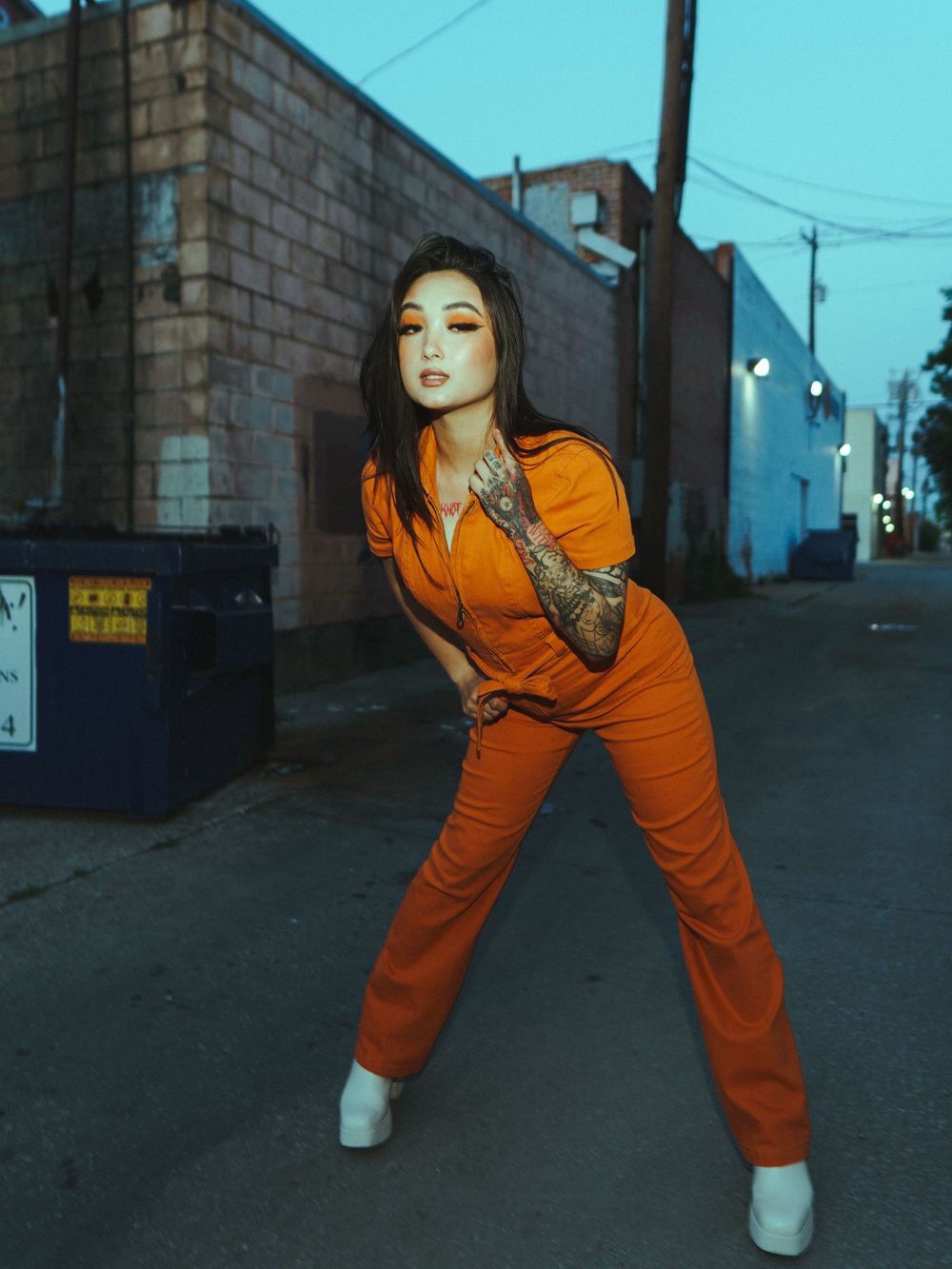 Stylish orange jumpsuit street fashion photography with dramatic dusk lighting and industrial backdrop.