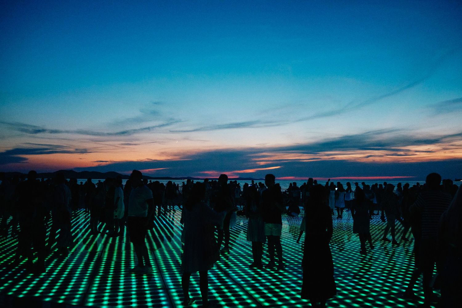 Crowd silhouettes stand on a glowing grid floor against a vibrant sunset sky.