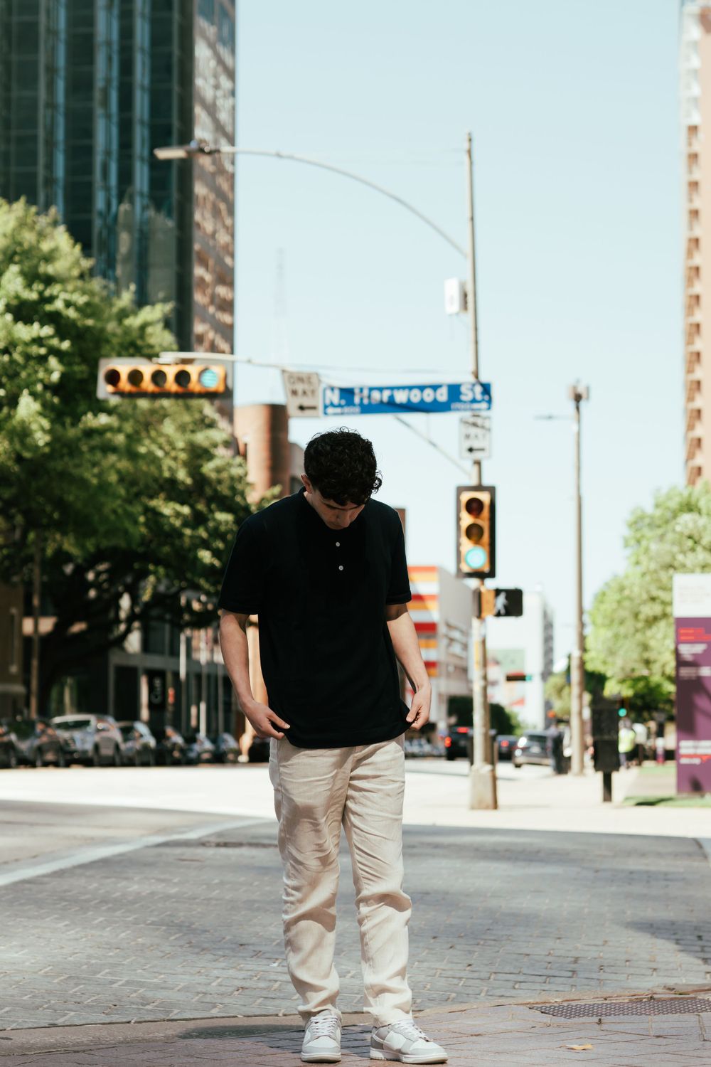 Person wearing black polo and khaki pants standing on an urban street corner with tall buildings in background.