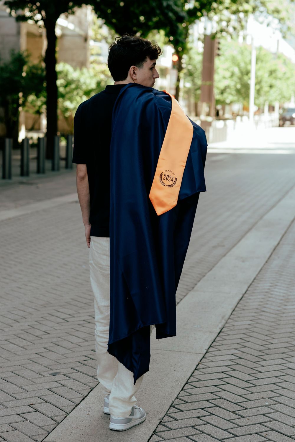 Graduate wearing black polo and graduation gown with orange stole walking on brick pathway.