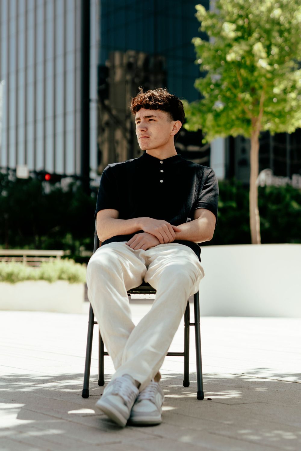 Student in black polo and khaki pants sitting casually on chair in urban setting with modern architecture.