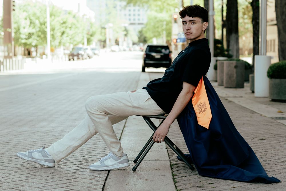 Graduate in commencement gown holding folding chair on brick sidewalk with orange graduation stole.