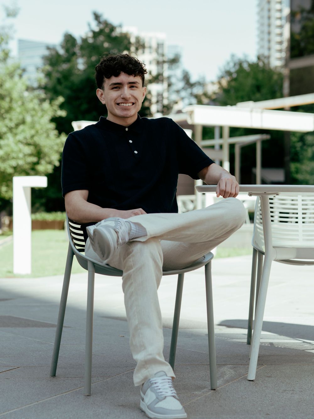 Student relaxing in white chair wearing black polo and khaki pants against urban backdrop.