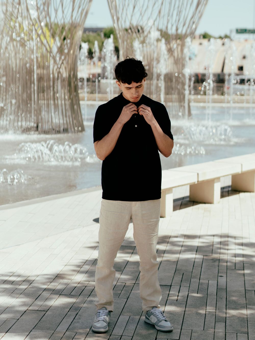 Person in casual attire standing by fountain adjusting shirt collar on sunny day.
