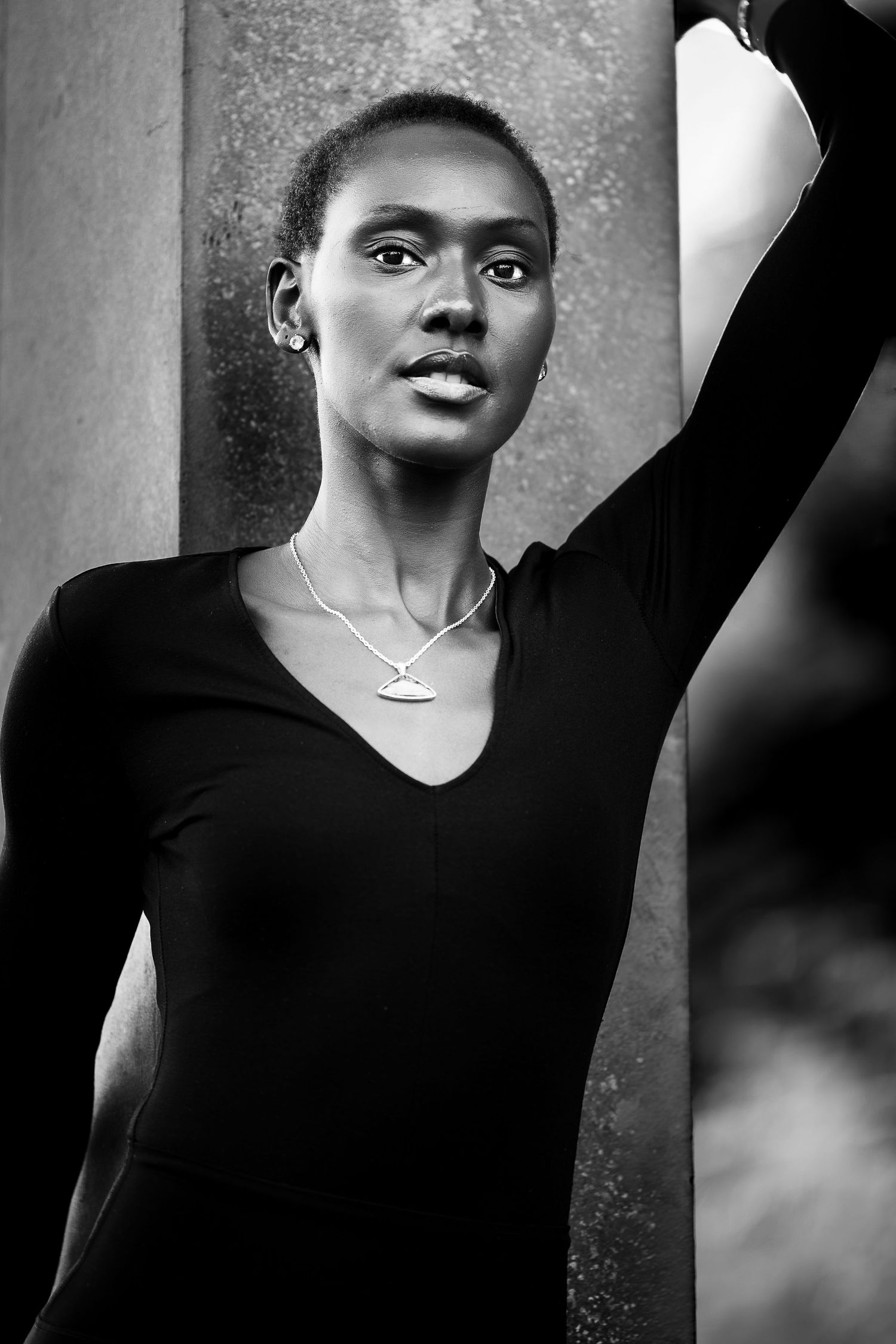 Black and white portrait of a woman in a black v-neck top and silver necklace, arm raised against a stone column.