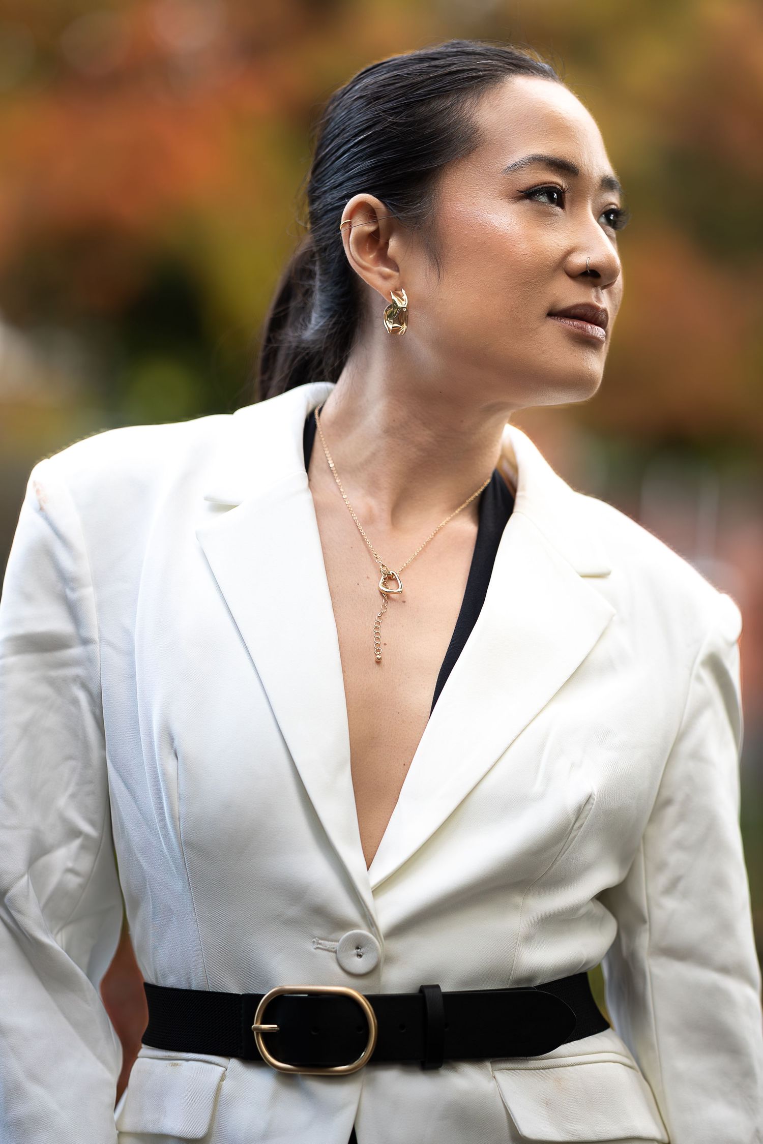 Woman in white blazer with black belt and gold necklace, looking sideways against autumn foliage background.