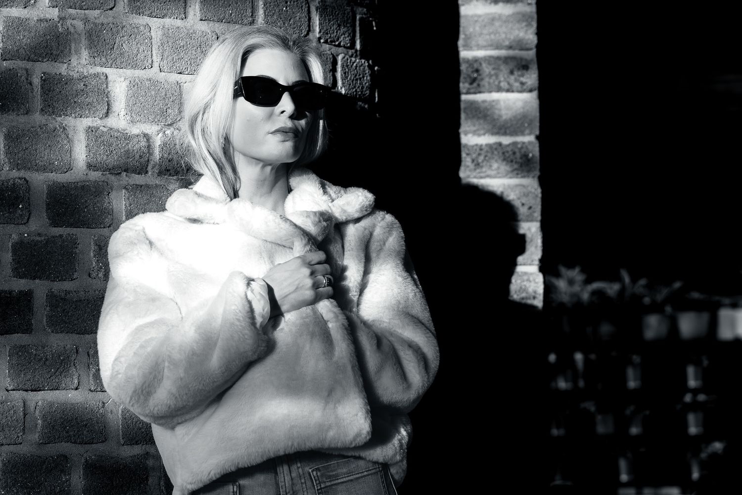 Black and white photo of someone wearing white coat and sunglasses leaning against brick wall in dramatic lighting.