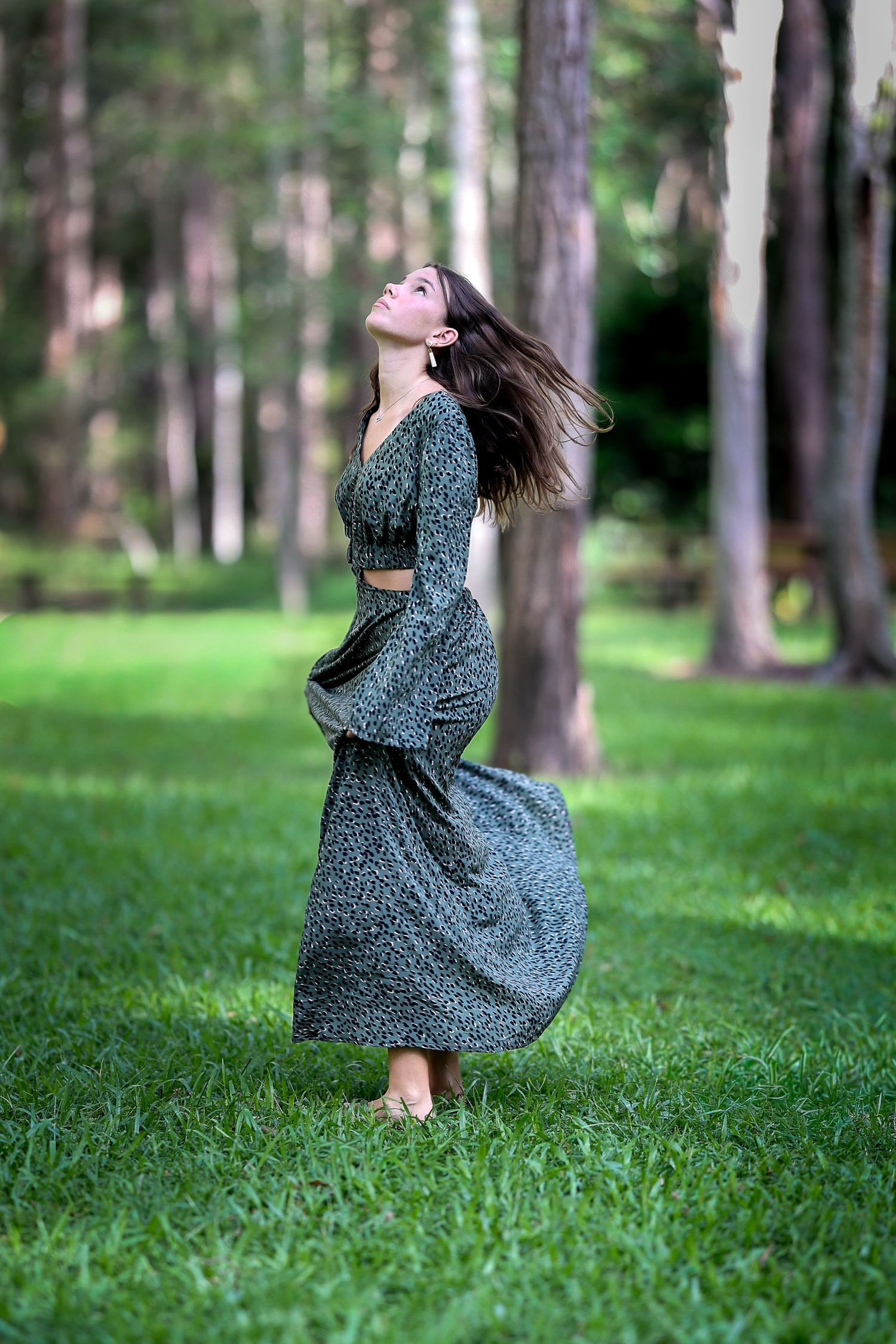 Woman in green floral maxi dress twirling in lush forest park, hair flowing, looking up joyfully.
