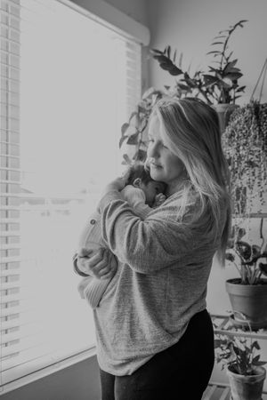 Black and white photo showing a person holding an infant near a window with houseplants in the background.