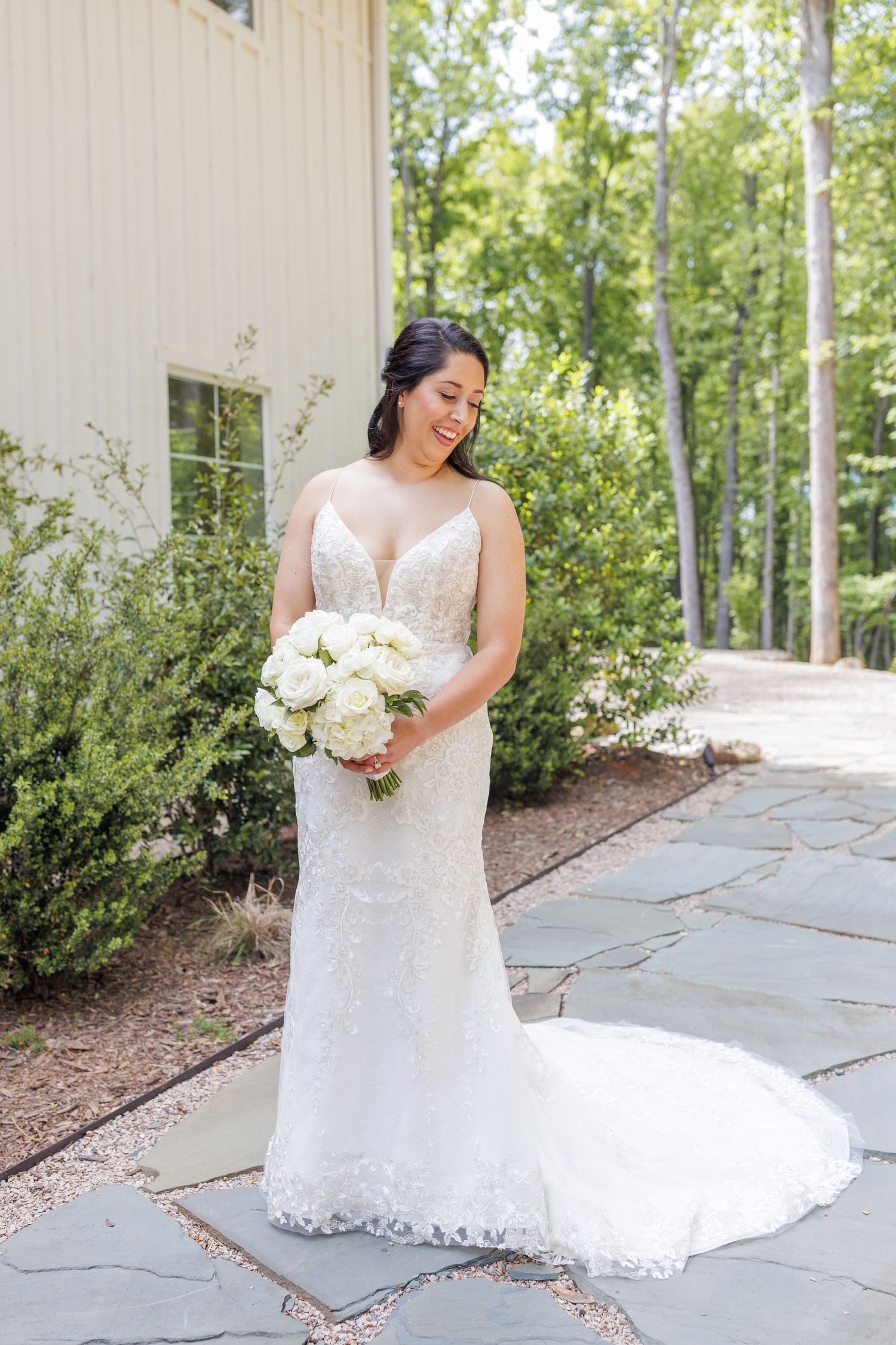 A bride in a fitted lace wedding gown holds a white bouquet while standing on a stone path surrounded by greenery at Carolina Grove in NC