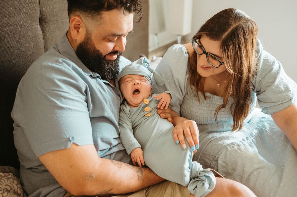 Happy new parents admire their yawning newborn baby dressed in a blue outfit and knit hat.