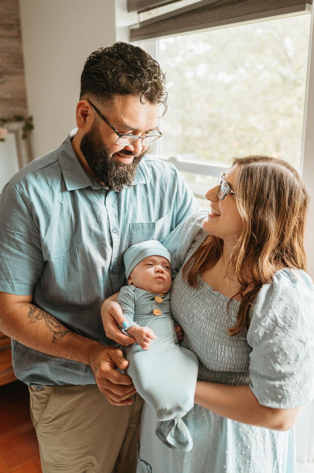 Smiling new parents in matching blue outfits hold their sleeping swaddled newborn by a bright window.