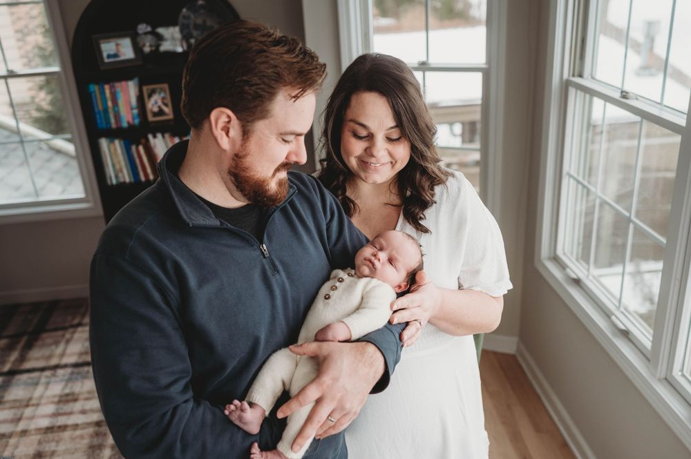 New parents smile lovingly at their newborn baby near bright windows in a lifestyle home portrait.