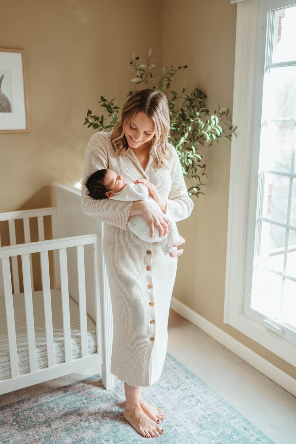 Smiling mother in cream button dress holding newborn baby in bright nursery by window.