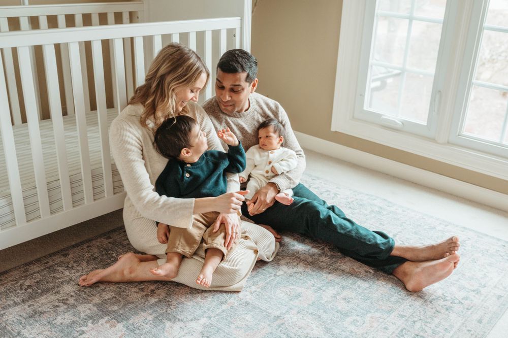 Young family sitting on nursery floor with toddler and newborn baby by white crib.