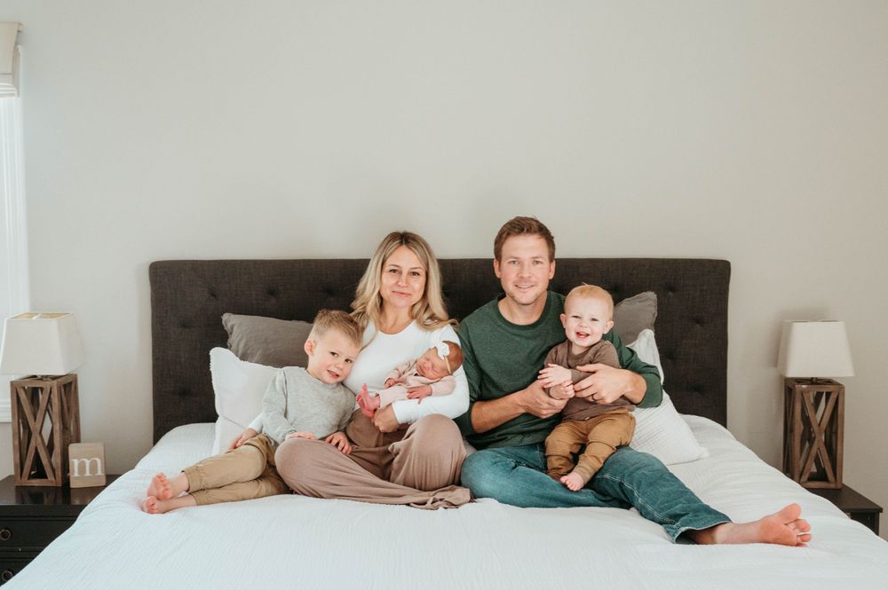 A cheerful family of five sits together on a white bed in a bright, cozy bedroom.