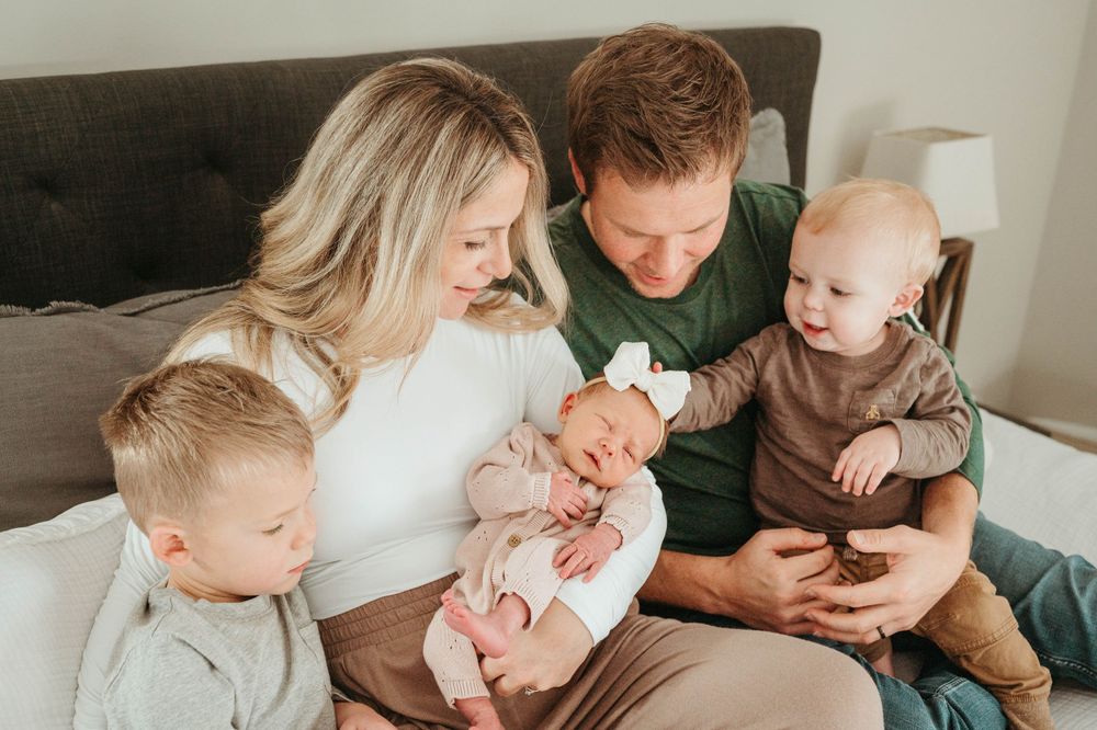 A family of five sits on a bed, parents lovingly holding their newborn and toddler children.