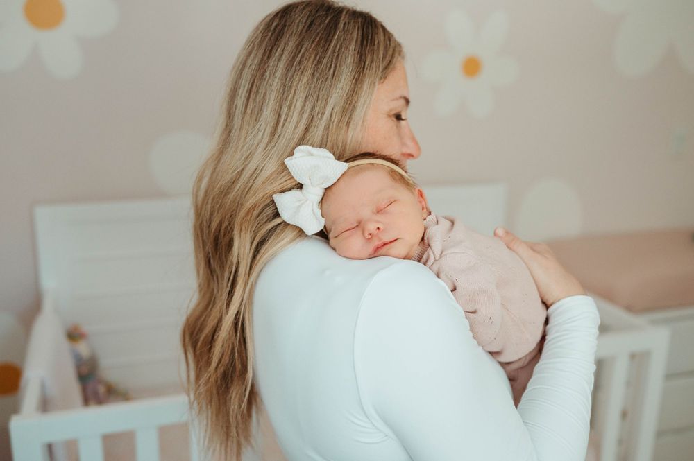 Mother with long blonde hair holds sleeping newborn in pink outfit against her shoulder in a soft nursery.