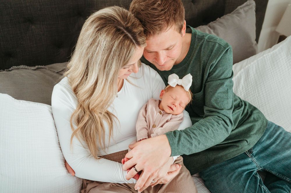 New parents in green and white outfits cuddle their sleeping newborn baby with a white bow headband.