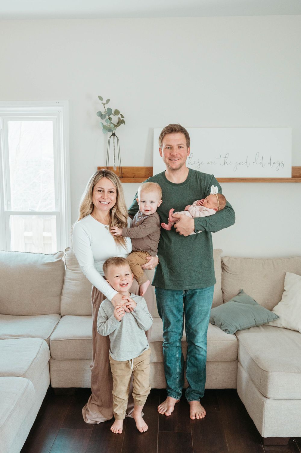 Smiling family of five with two toddlers and a newborn poses together in a bright modern living room.