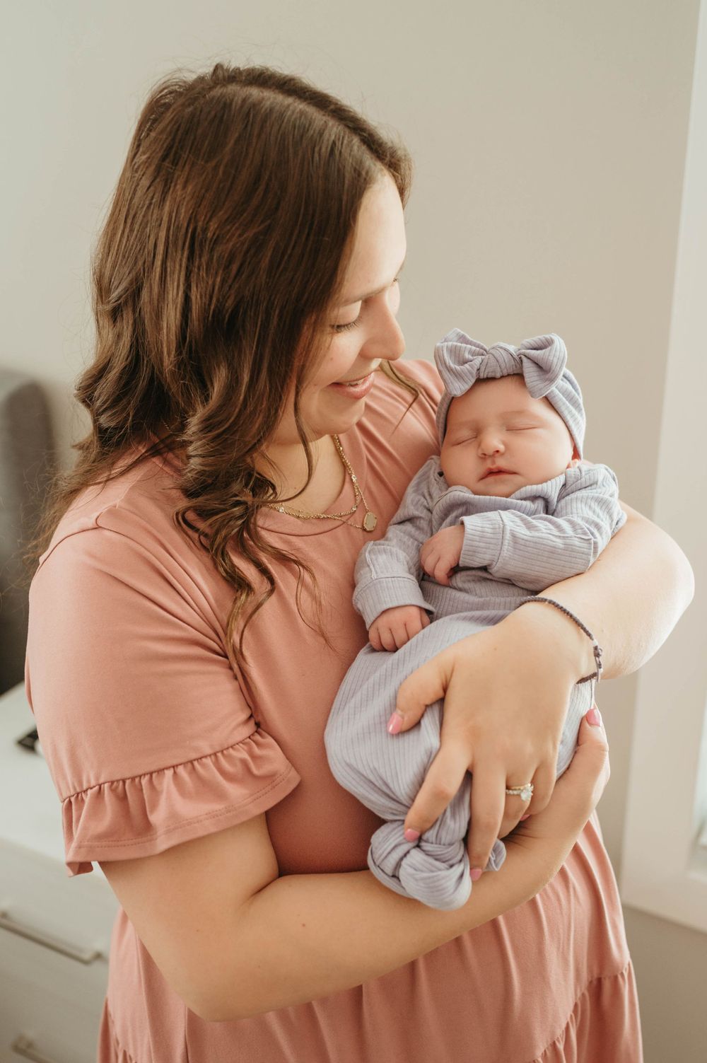 Brunette mother in pink dress cradles her sleeping newborn baby girl in a gray bow headband.