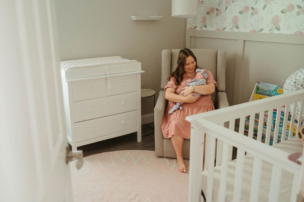 Mother sits in nursery glider chair cradling her newborn baby surrounded by white furniture.