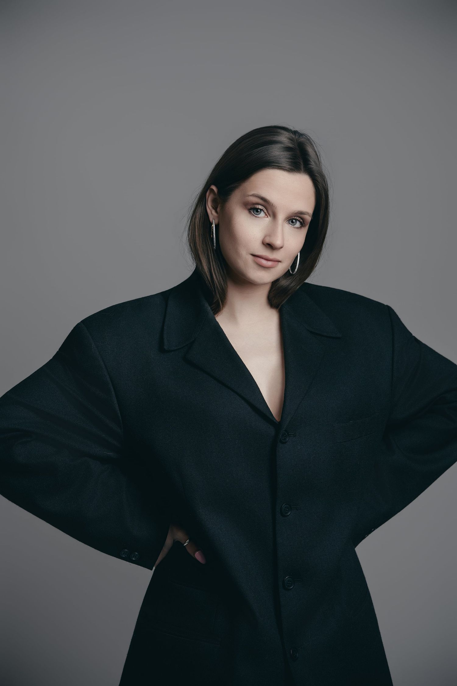 Young woman in oversized black blazer posing confidently against a neutral gray studio background.