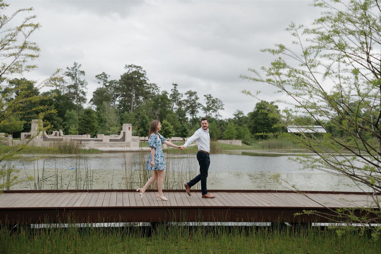A couple stands together on a wooden dock overlooking a serene lake surrounded by trees on an overcast day.