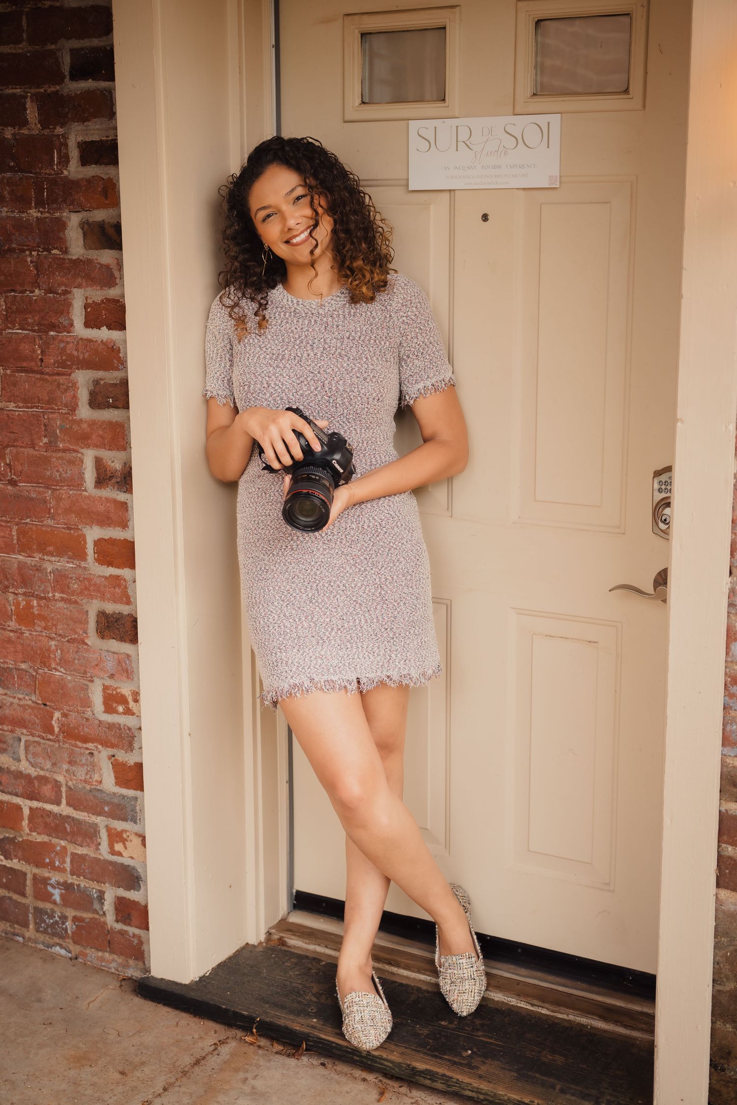 Photographer in gray dress posing creatively in front of a beige door.