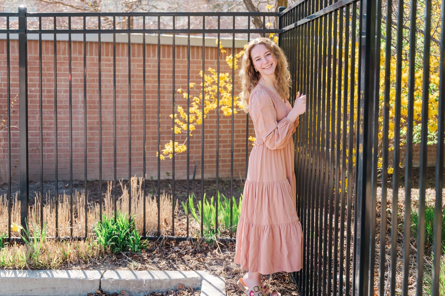 Woman in maxi dress poses against iron gate with spring garden backdrop.