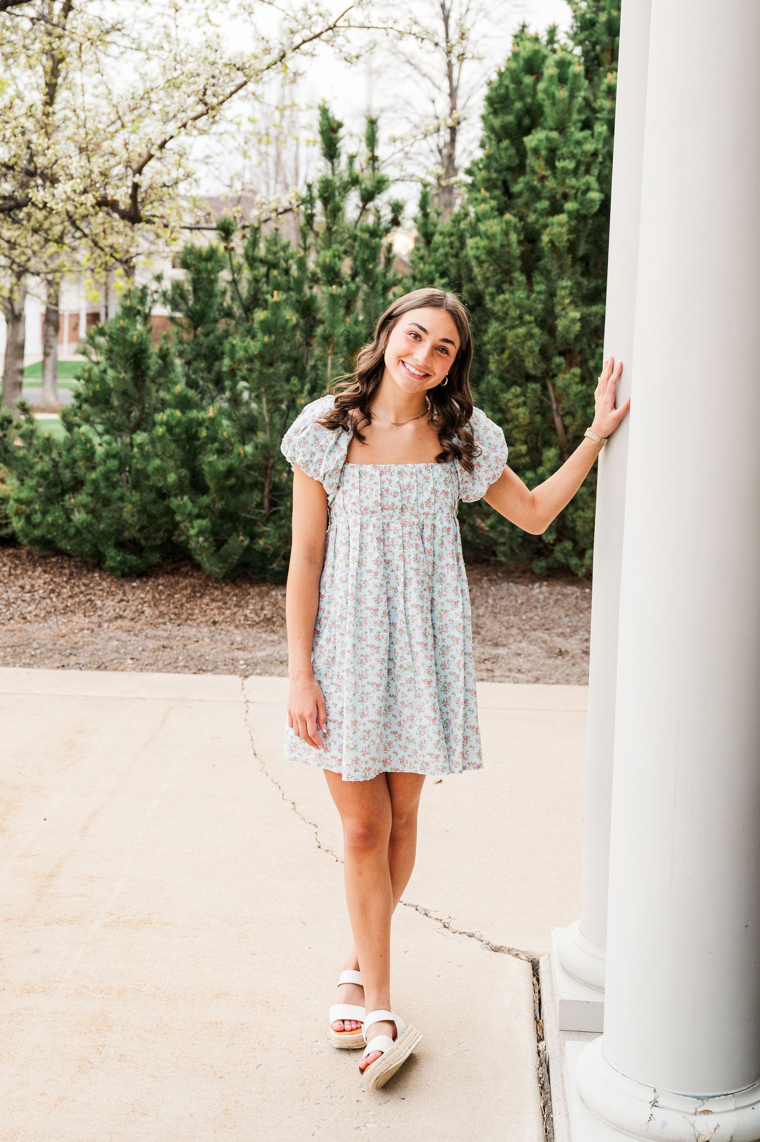 Woman in floral dress posing by white column with evergreen trees in background.
