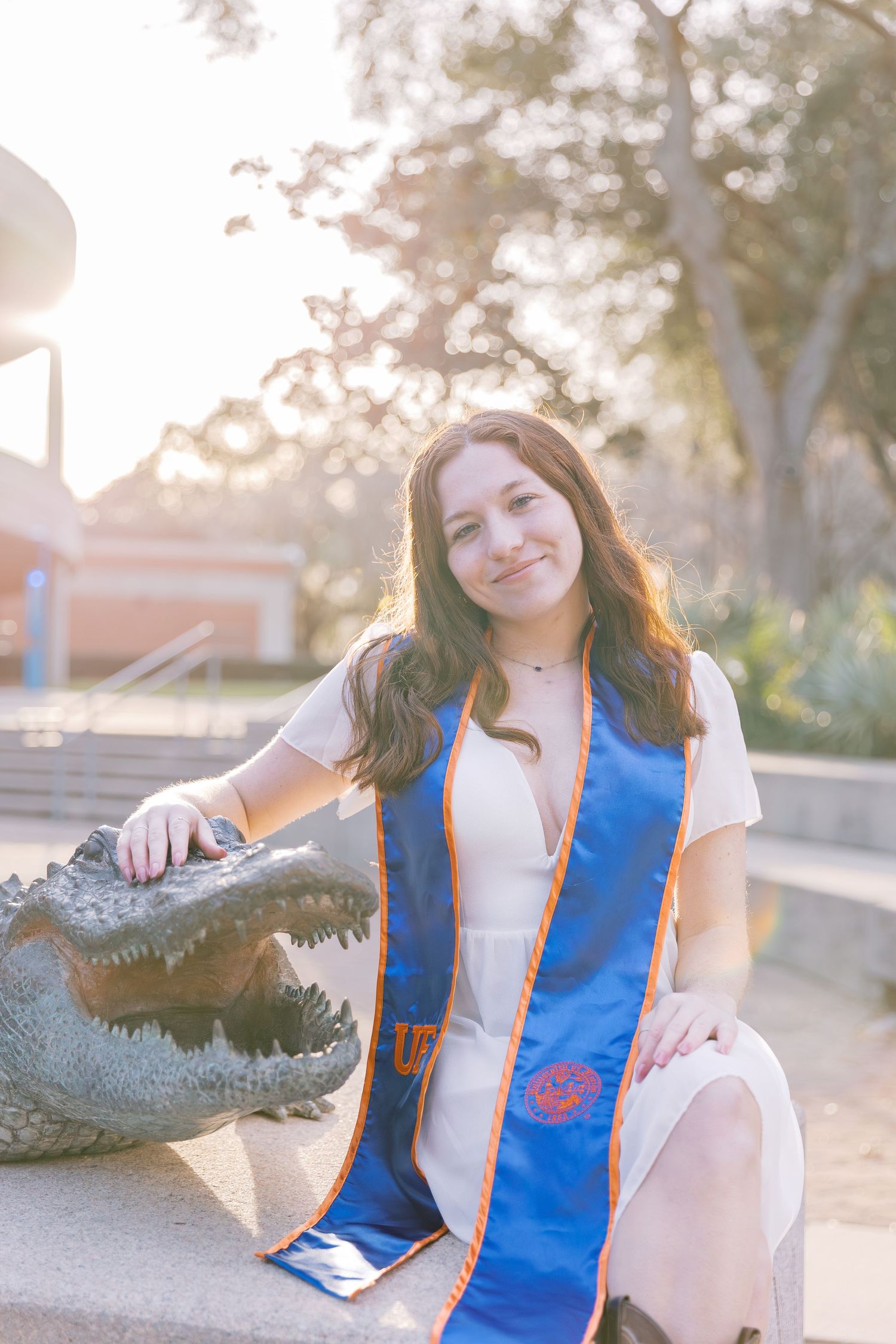 Graduate in white dress and blue sash poses with alligator statue during golden hour.