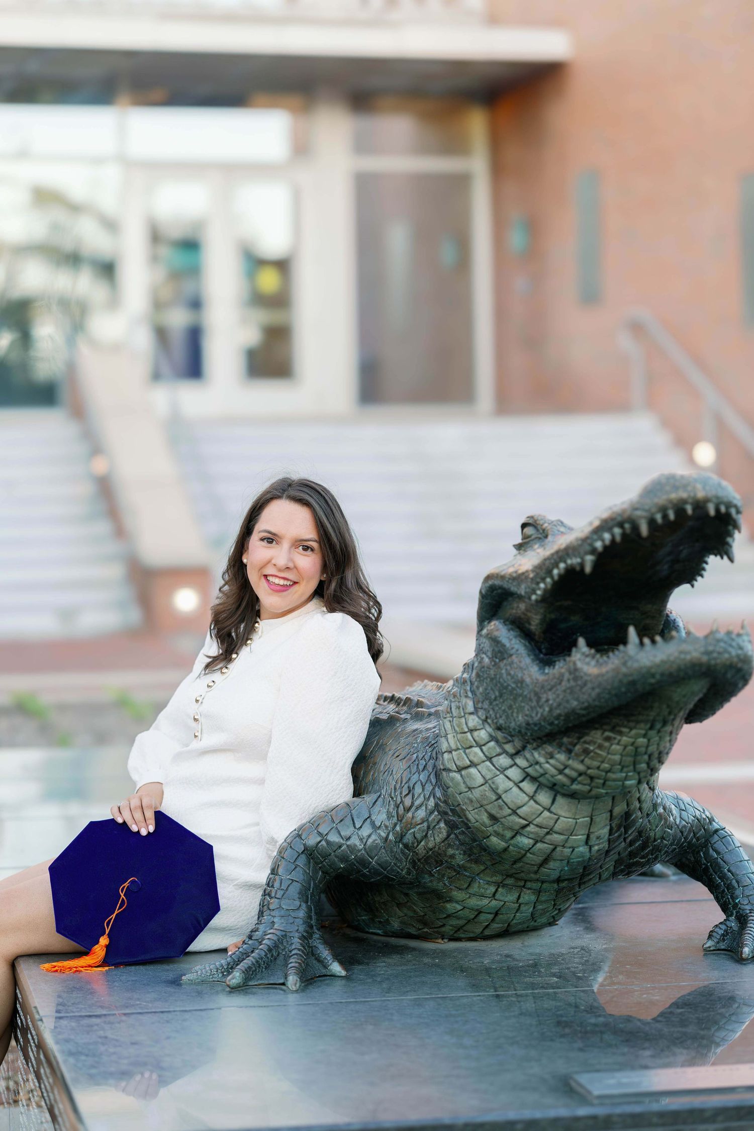 Graduate in white dress and blue sash poses with alligator statue during golden hour.