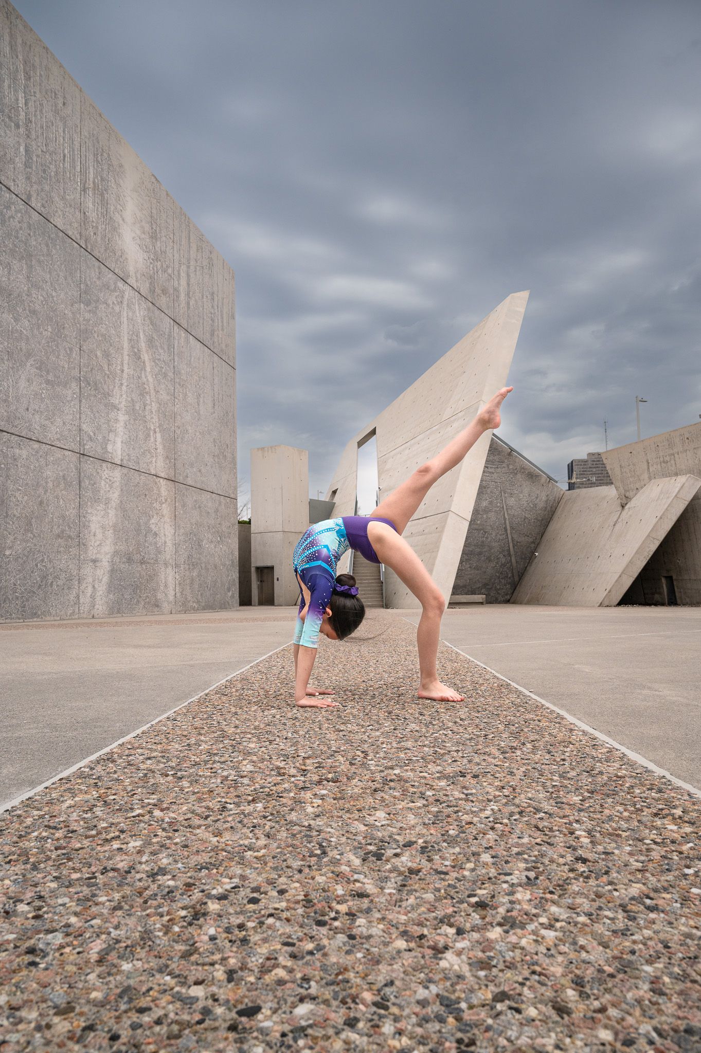 Gymnast performs handstand on textured ground against angular concrete structure.