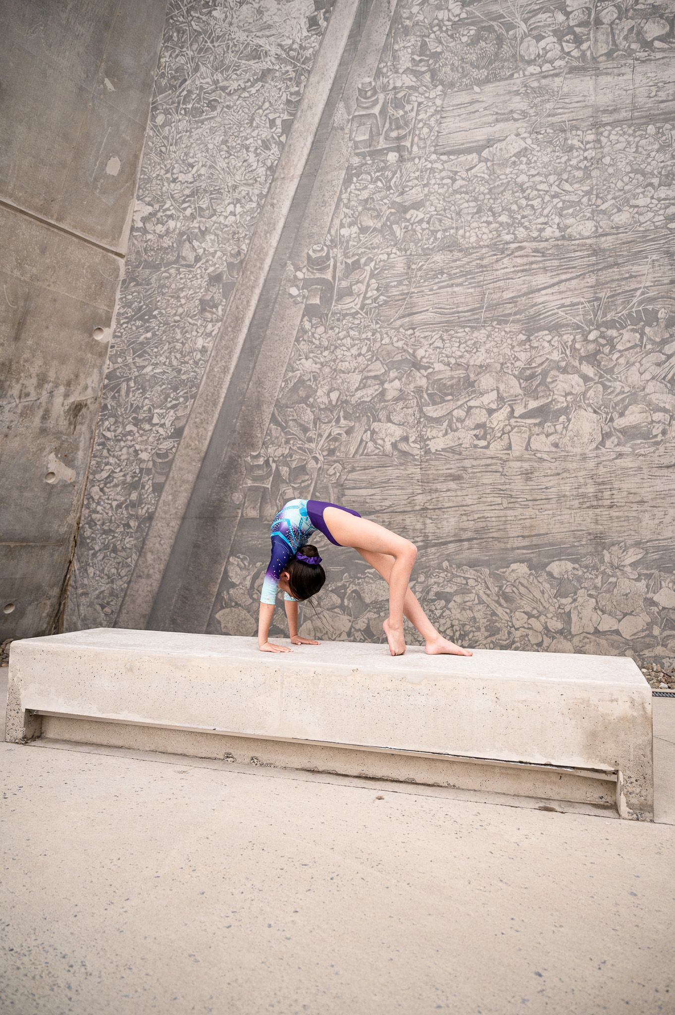 Gymnast performing backbend on concrete bench against textured wall.