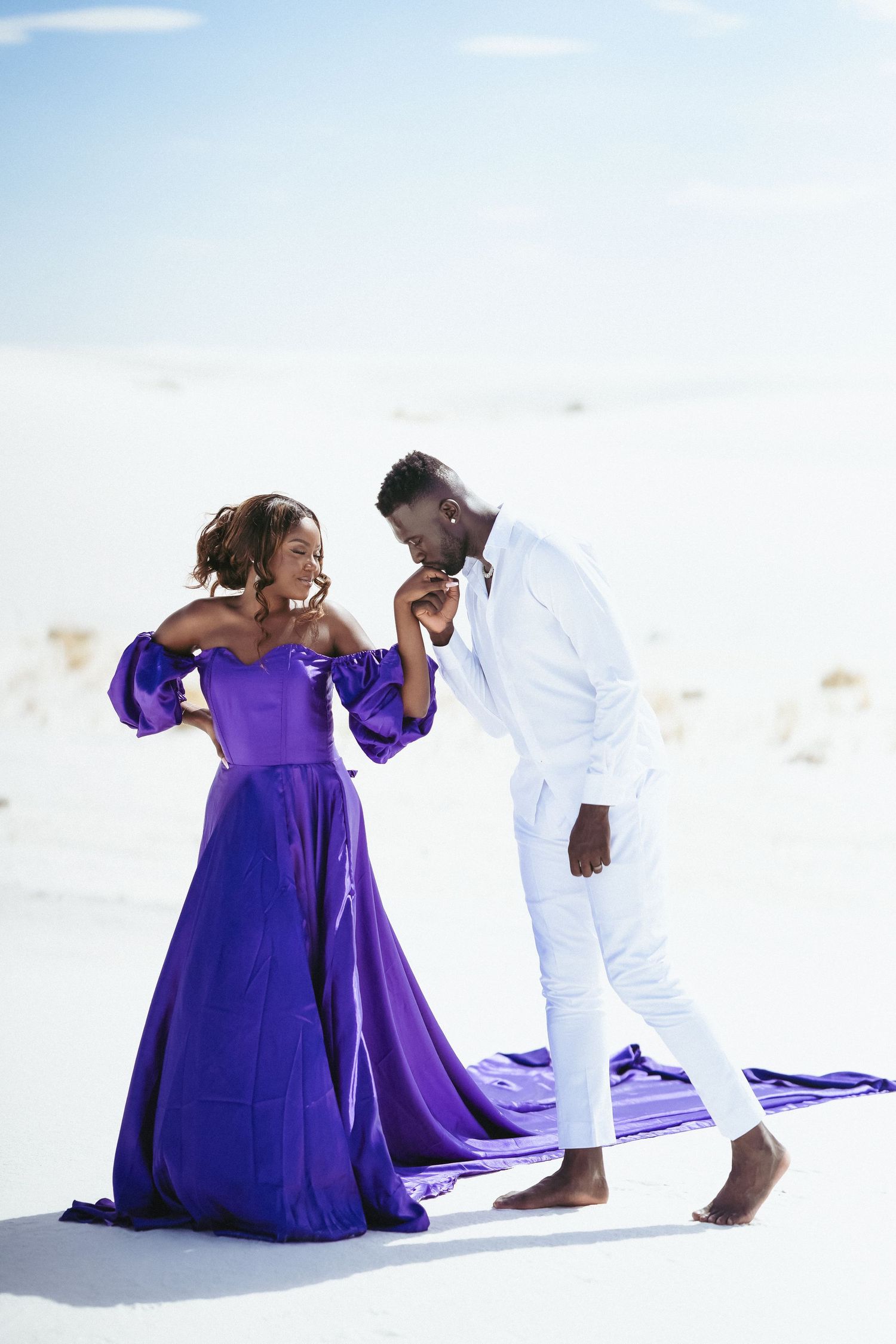 Couple in purple dress and white outfit shares tender moment on sand dunes.
