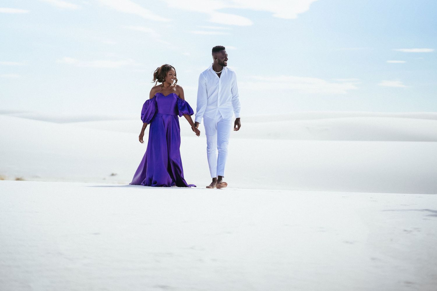 Two figures walk together across bright white sand dunes under blue sky.