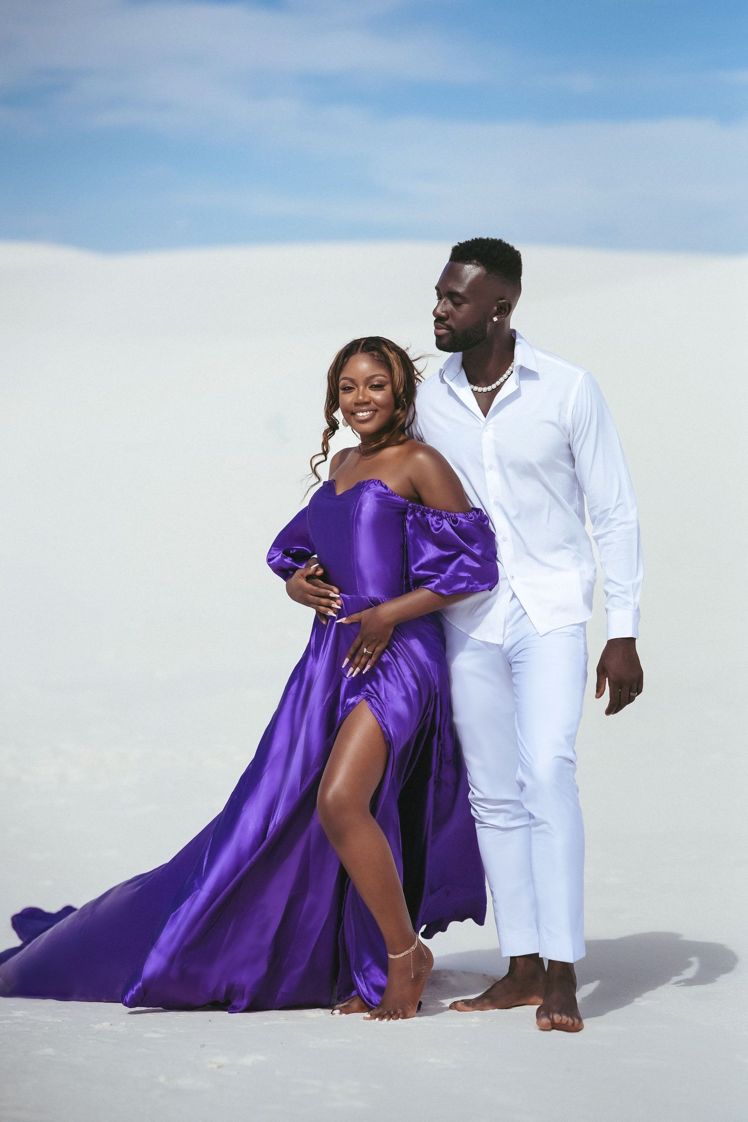 A couple in purple gown and white suit pose elegantly on white sand dunes.