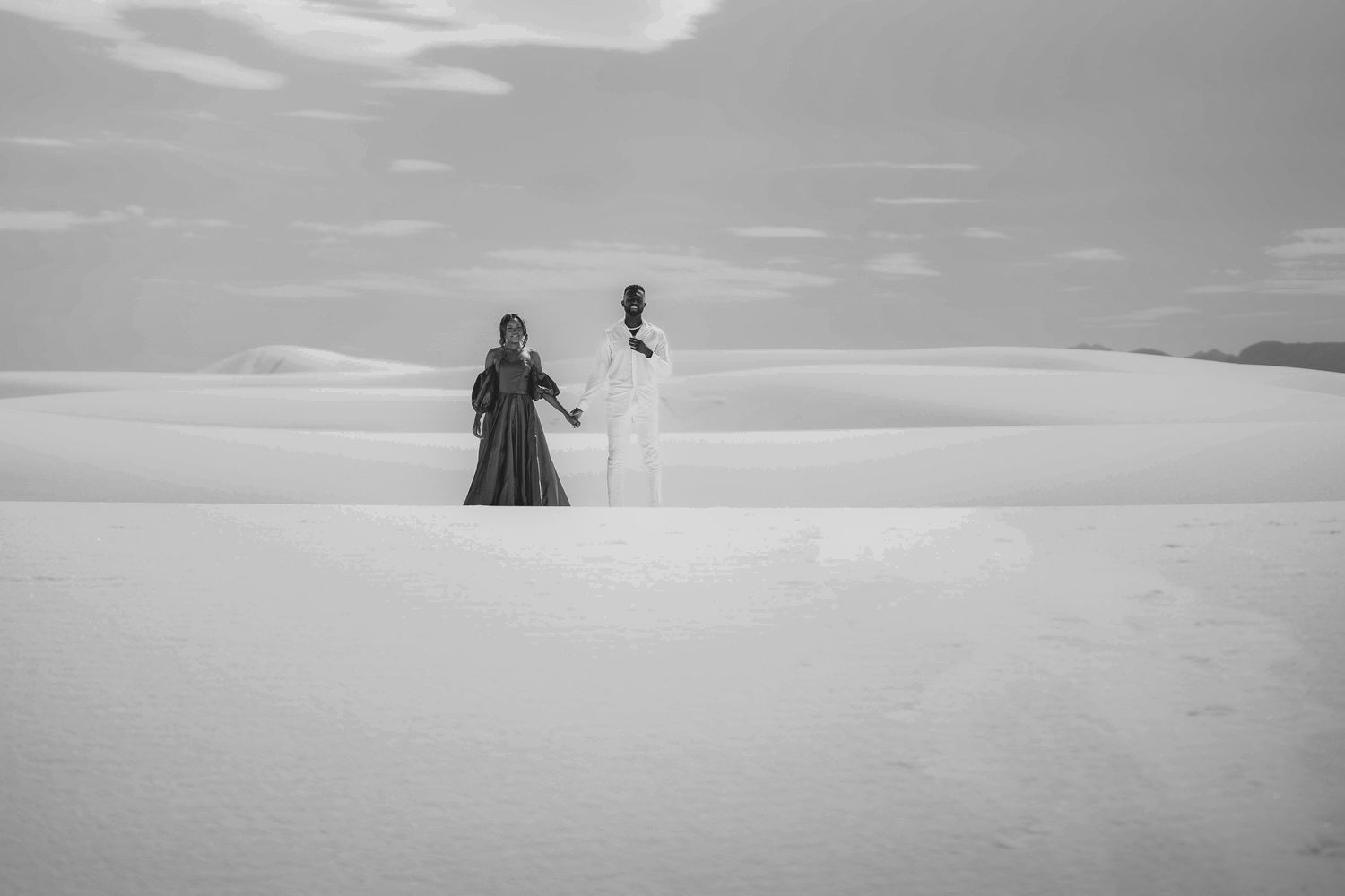 A black and white photo of two people standing on sand dunes under cloudy skies.