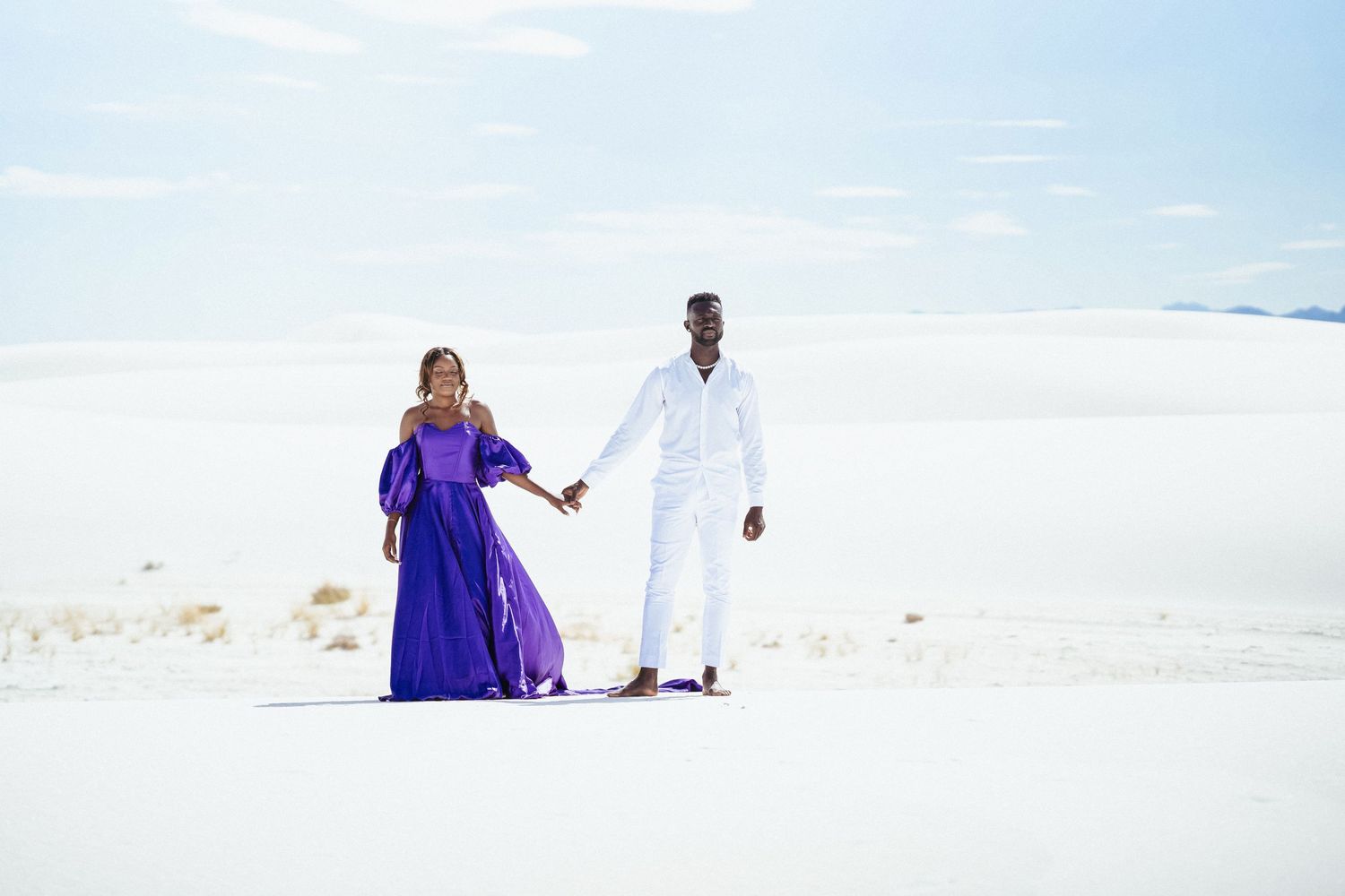 A couple in purple dress and white outfit holding hands while walking on white sand.
