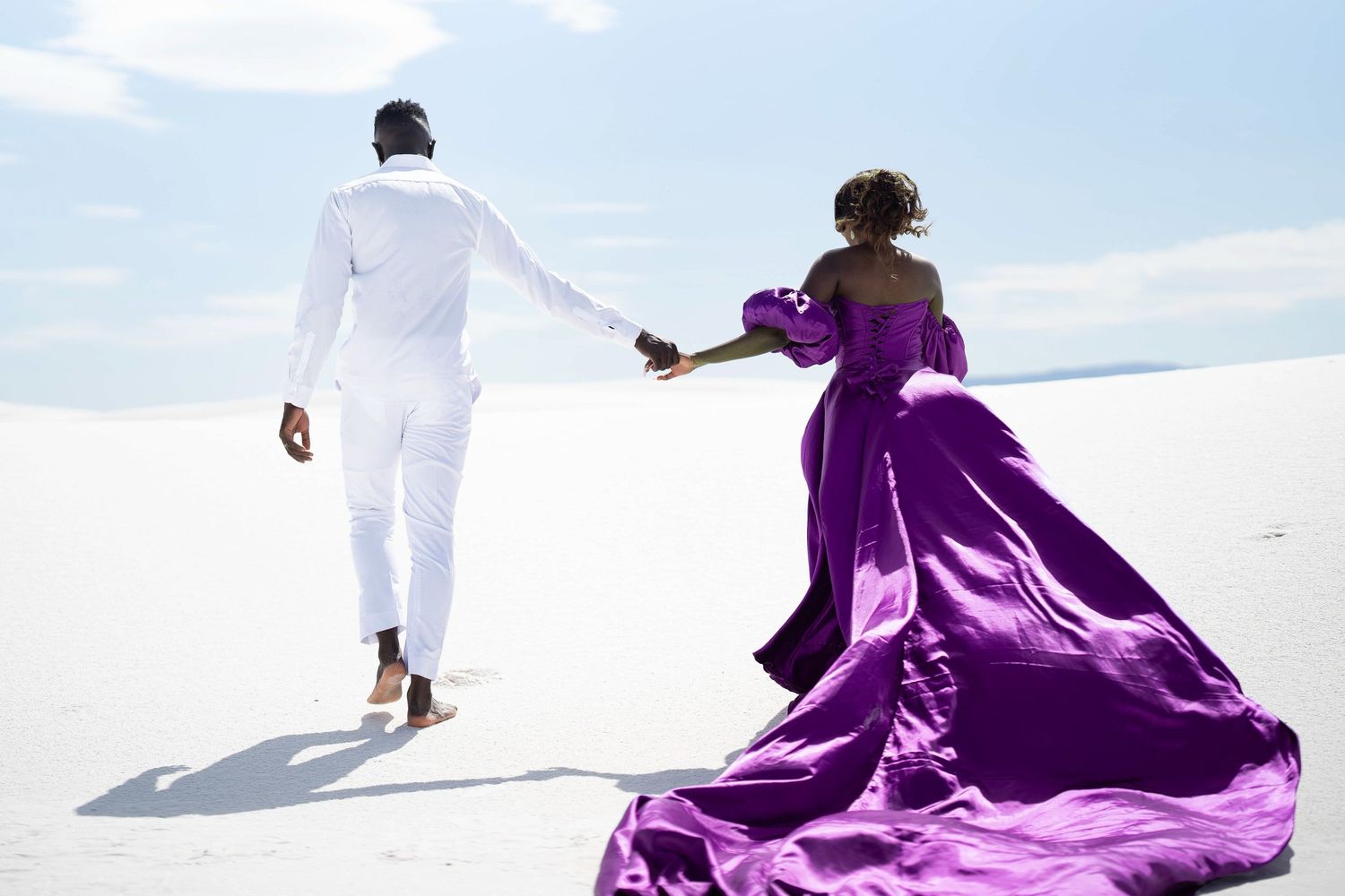 A couple poses dramatically in flowing purple and white outfits against white sand dunes under a bright blue sky.