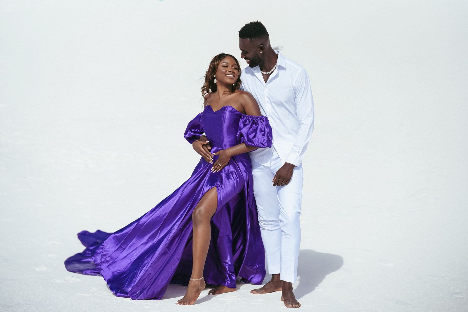 A couple in purple dress and white outfit pose romantically on a beach setting.